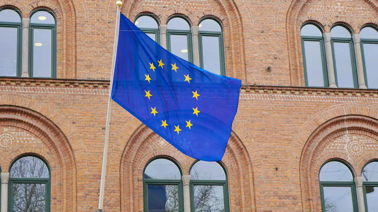 European Union flag with yellow stars flying in front of a brick building with arched windows.