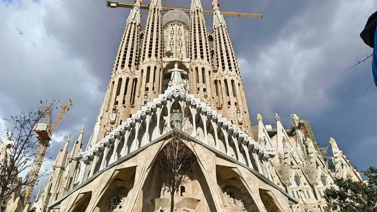 A low-angle view of the Sagrada Família in Barcelona, showcasing its intricate stone carvings, towering spires, and ongoing construction with visible cranes against a cloudy sky.