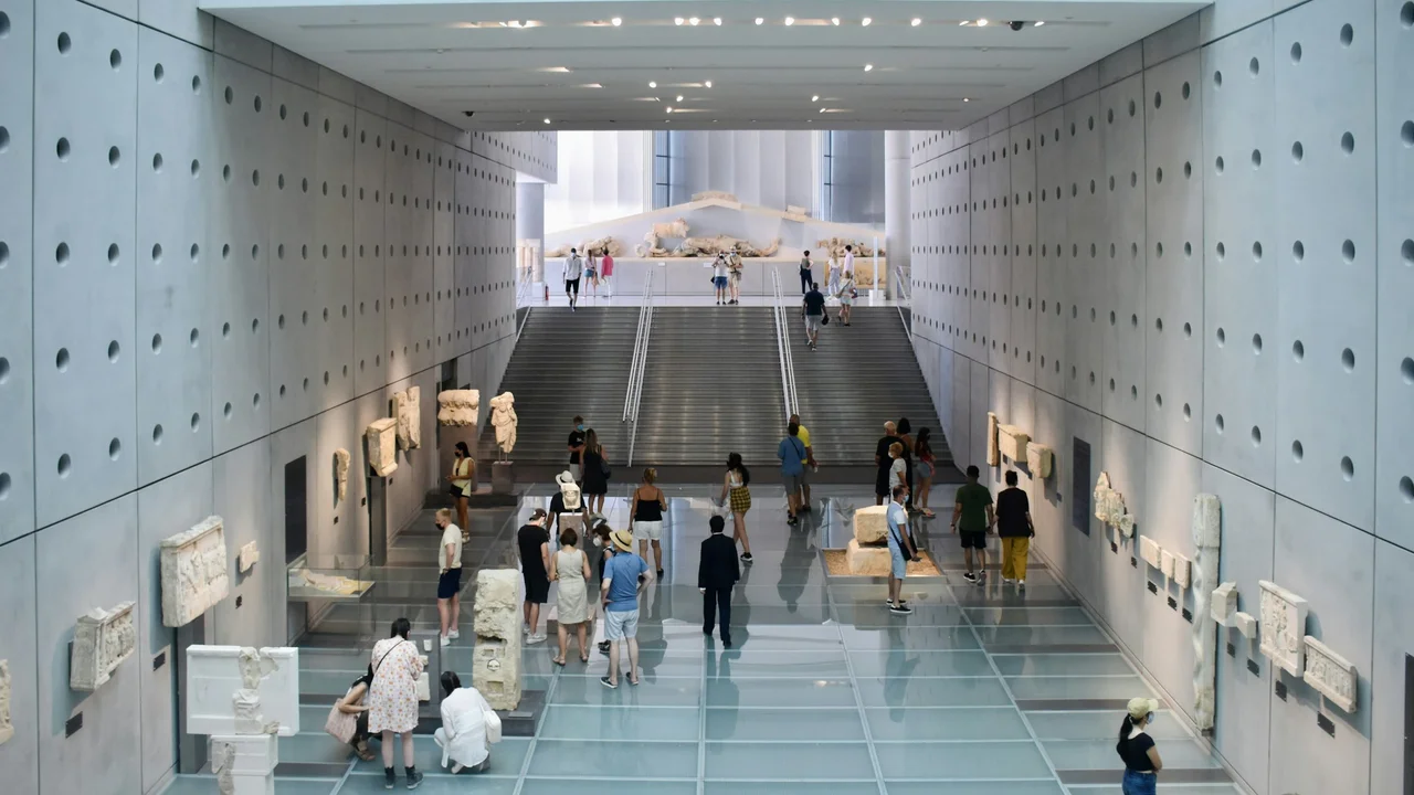 Interior of the Acropolis Museum gallery with modern concrete walls, glass floor, and visitors viewing ancient Greek marble sculptures and architectural fragments beneath a wide staircase.