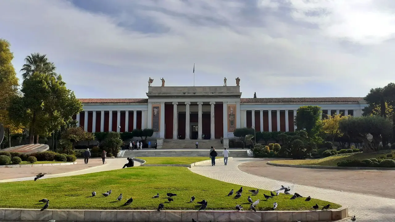 Exterior of the National Archaeological Museum of Athens, featuring a neoclassical facade with columns, statues on the roof, landscaped gardens, and visitors walking across the front lawn.
