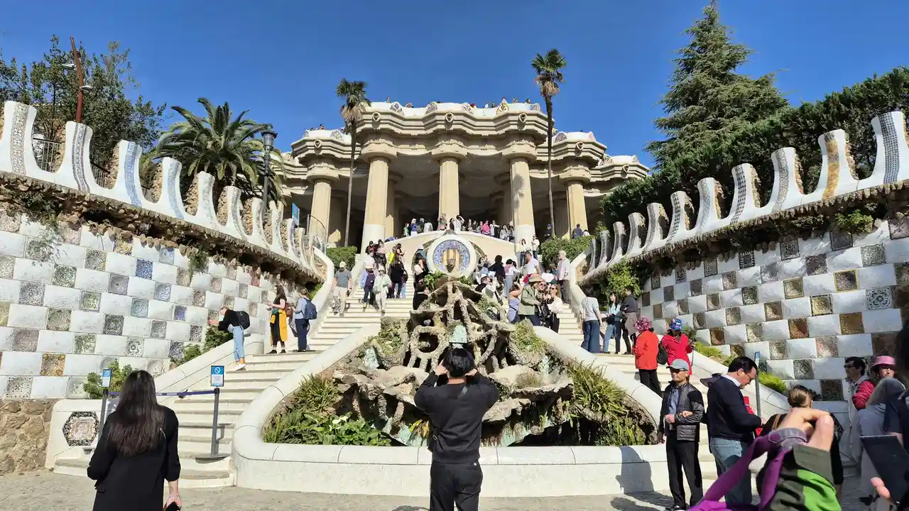 A wide-angle shot of the grand staircase at Park Güell in Barcelona, featuring colorful mosaic walls, a central fountain, and the iconic Hypostyle Room with large stone columns in the background under a clear blue sky.
