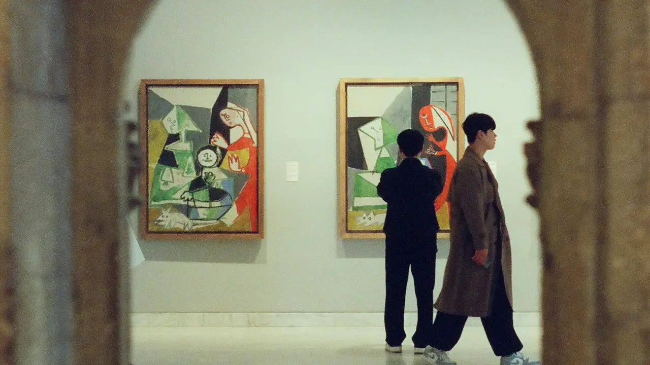 A view through a stone archway into a gallery at the Picasso Museum, where two visitors are observing two large cubist paintings featuring abstract figures and bold colors hanging on a white wall.