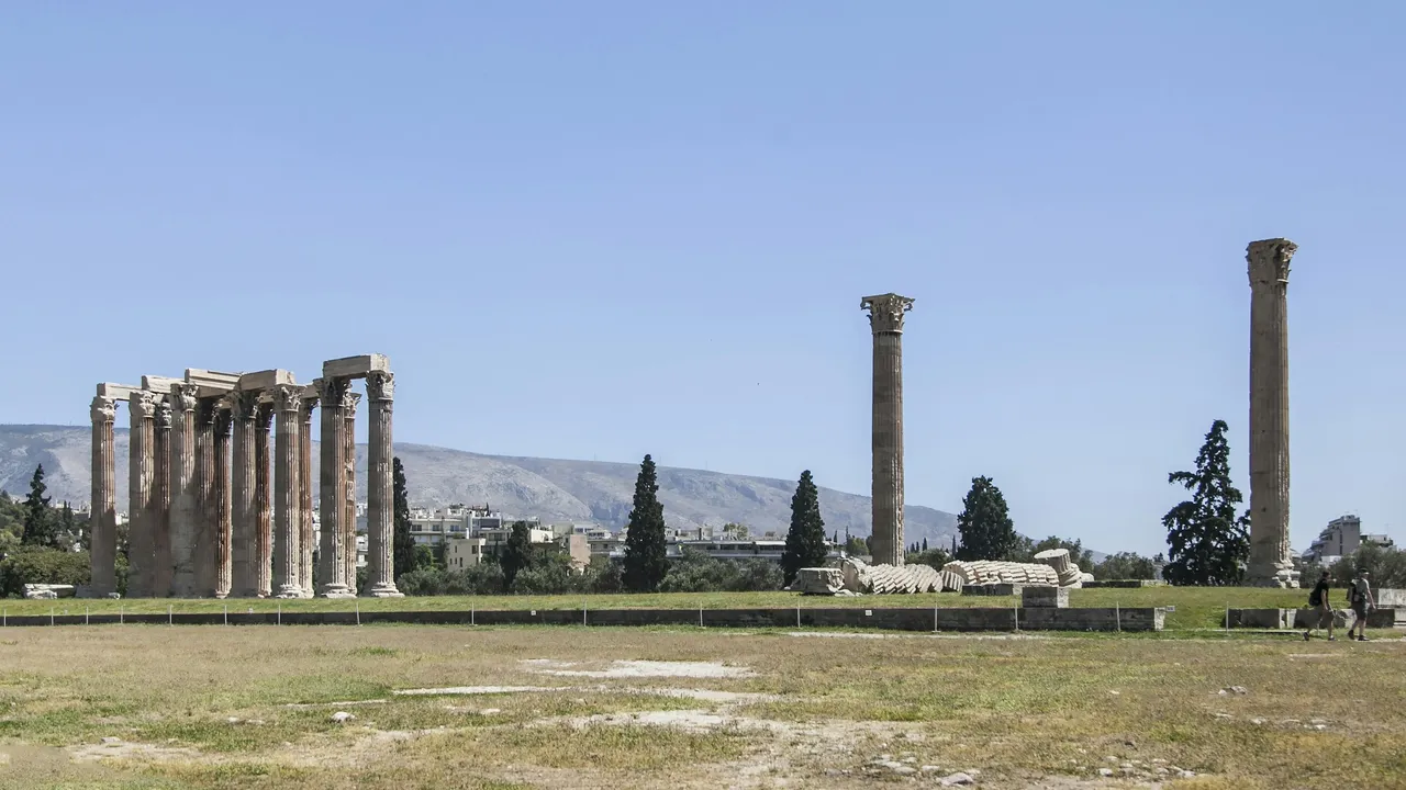 Ruins of the Temple of Olympian Zeus in Athens, with towering Corinthian columns standing in an open field against a clear blue sky and city buildings in the background.