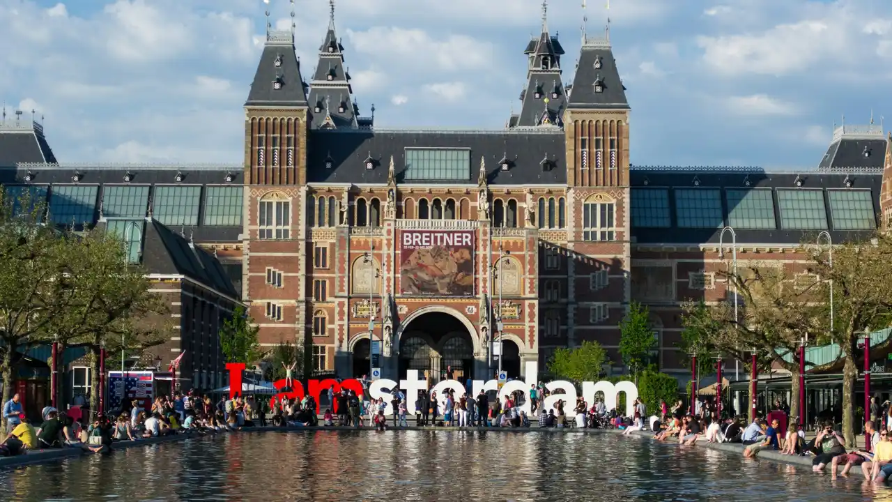 Amsterdam Museum exterior at Museum Square featuring historic Dutch architecture, the iconic “I Amsterdam” sign, and visitors relaxing by the reflecting pool, highlighting Dutch history and cultural heritage in Amsterdam.