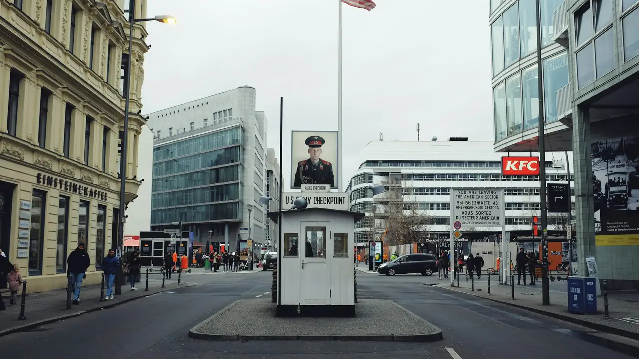 Historic Checkpoint Charlie in Berlin featuring the iconic guardhouse, Cold War signage, surrounding city streets, and visitors exploring this famous former border crossing.