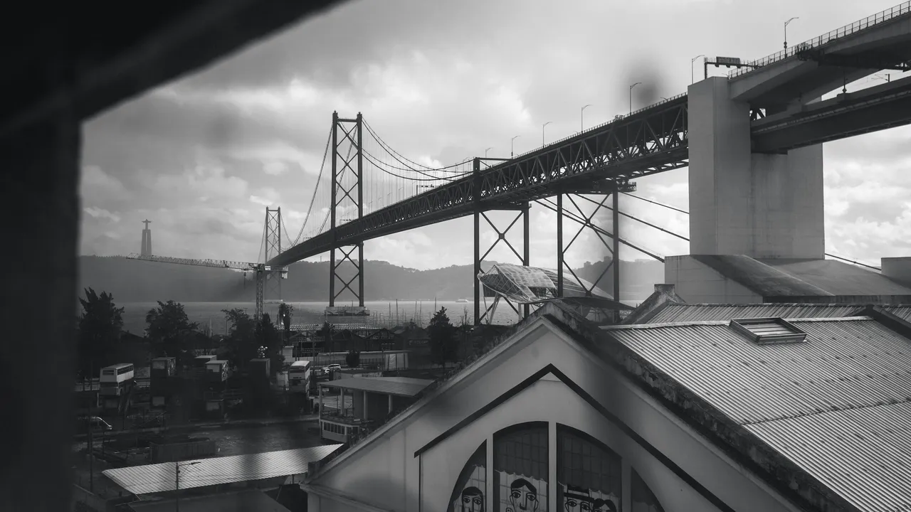 Black and white view of the 25 de Abril Bridge seen from LX Factory in Lisbon, featuring industrial rooftops, urban architecture, and the Tagus River under a dramatic cloudy sky.