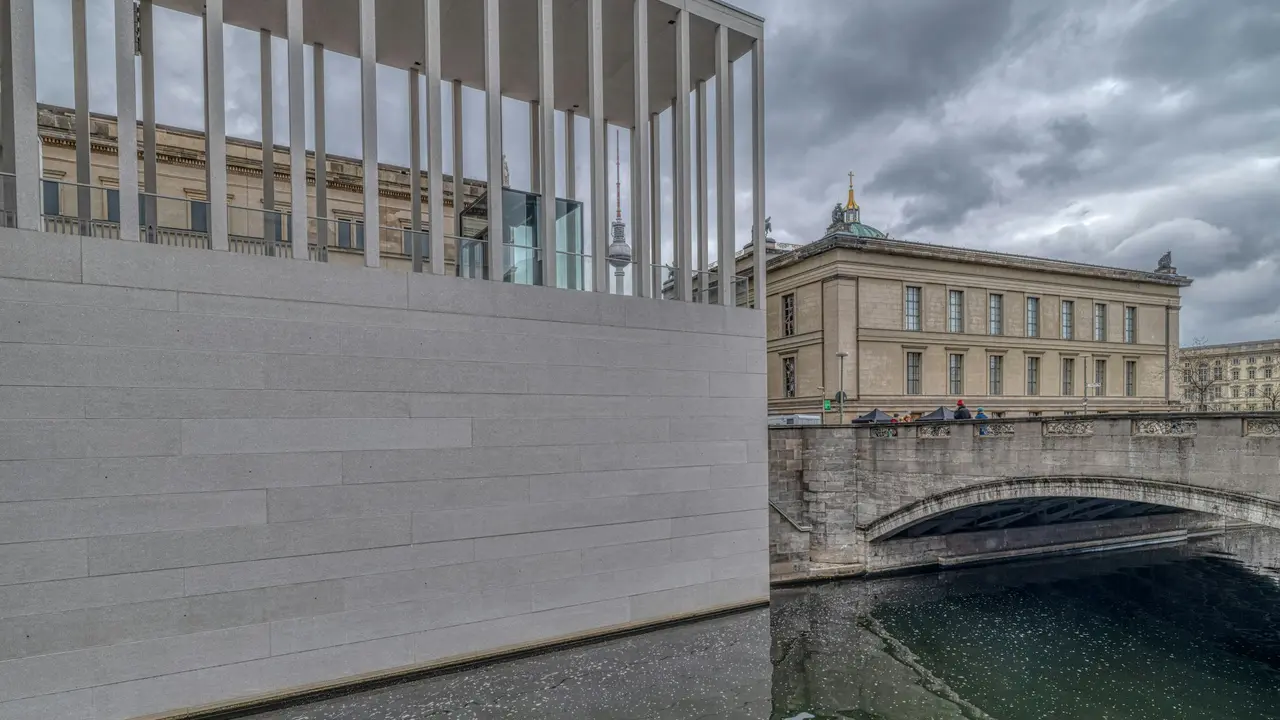 Modern James-Simon-Galerie entrance on Museum Island in Berlin with the Pergamon Museum and Spree River in the background, showcasing historic architecture under a cloudy sky.