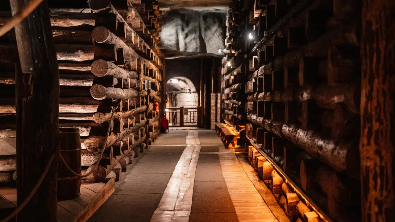 Underground salt mine tunnel with stacked timber supports and wooden beams lining both sides, illuminated walkway leading through historic mining corridor with warm industrial lighting.