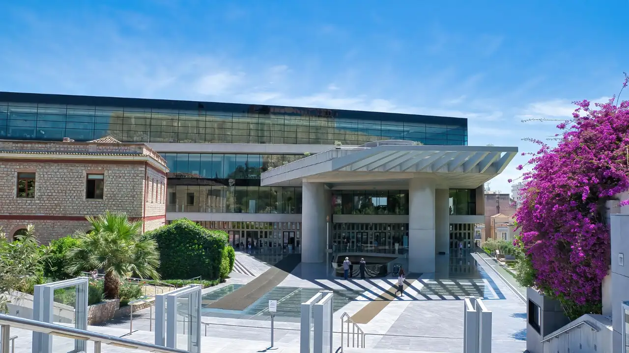 Modern contemporary museum building with glass façade and grand entrance canopy, surrounded by palm trees and vibrant purple bougainvillea under a clear blue sky.