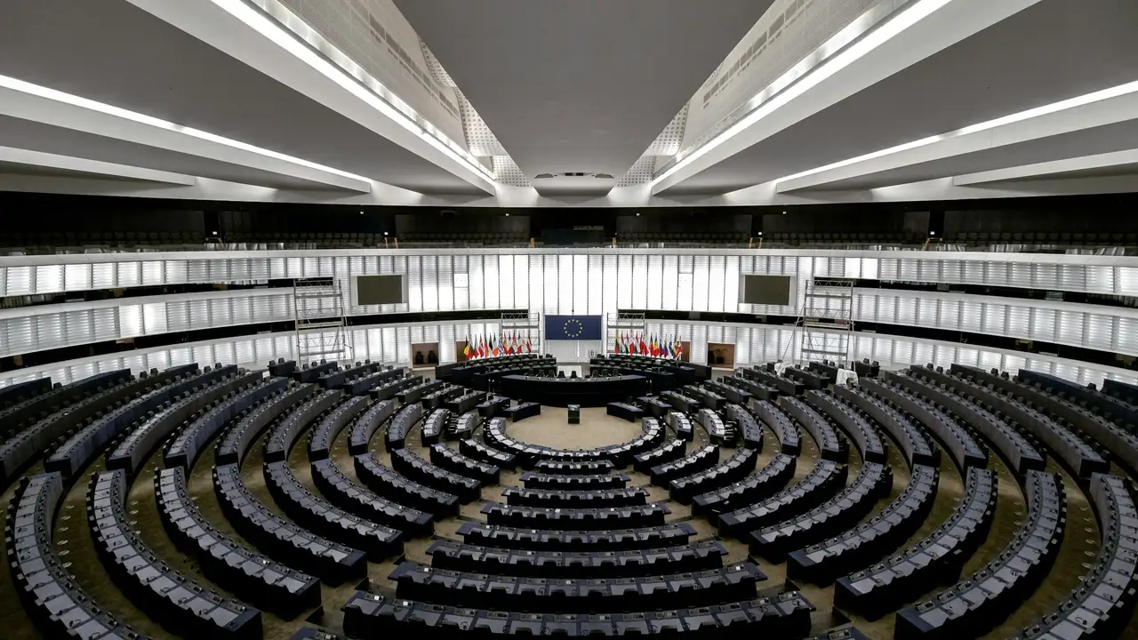Interior of the European Parliament chamber in Strasbourg with semicircular seating rows, EU flag and member state flags at the central podium, modern architecture and plenary hall setting