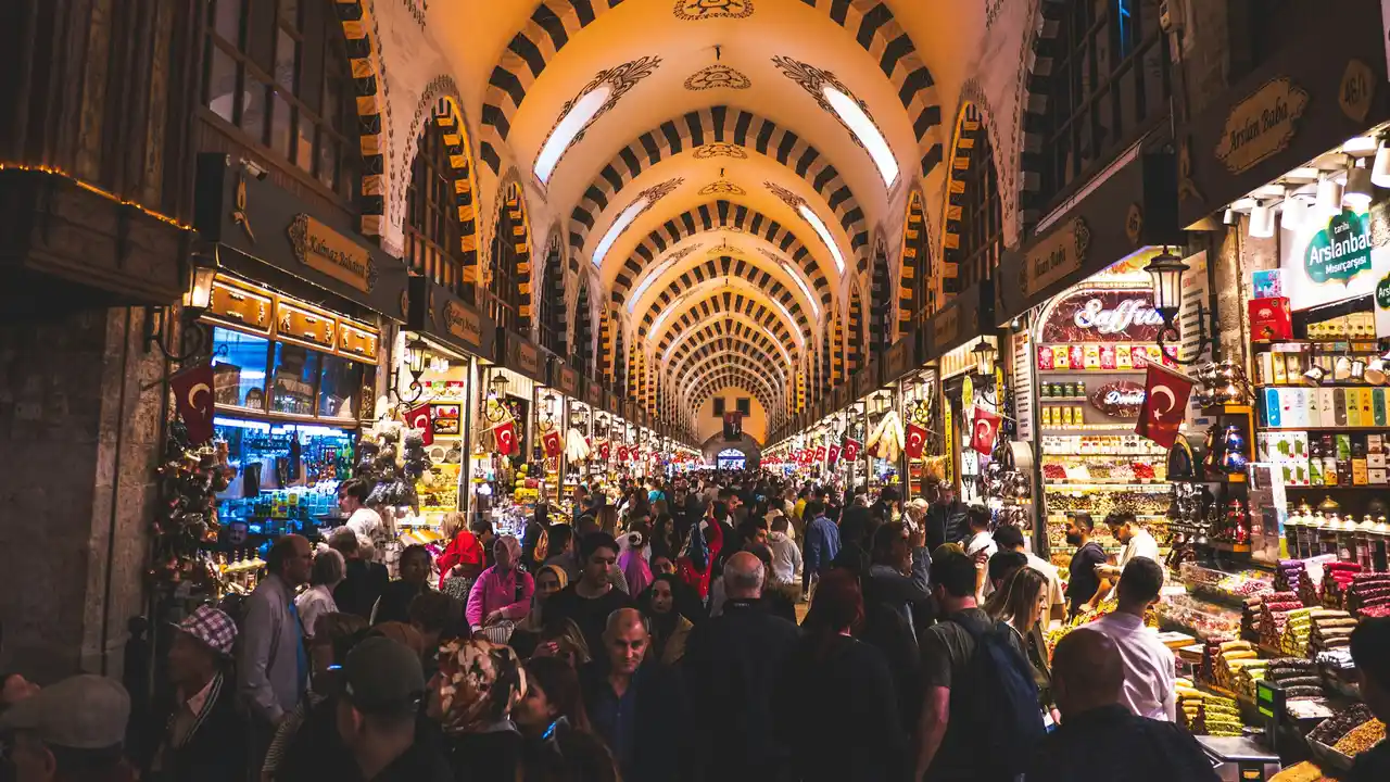 Crowded interior of the Grand Bazaar in Istanbul, Turkey, featuring vibrant market stalls, arched Ottoman ceilings, colorful lights, Turkish flags, and shoppers exploring traditional souvenirs and spices