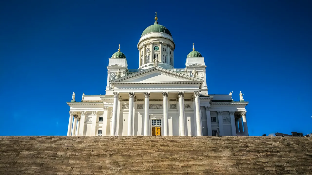 Helsinki Cathedral (Helsingin tuomiokirkko) with white neoclassical facade and green domes, viewed from Senate Square steps under a clear blue sky in Helsinki, Finland