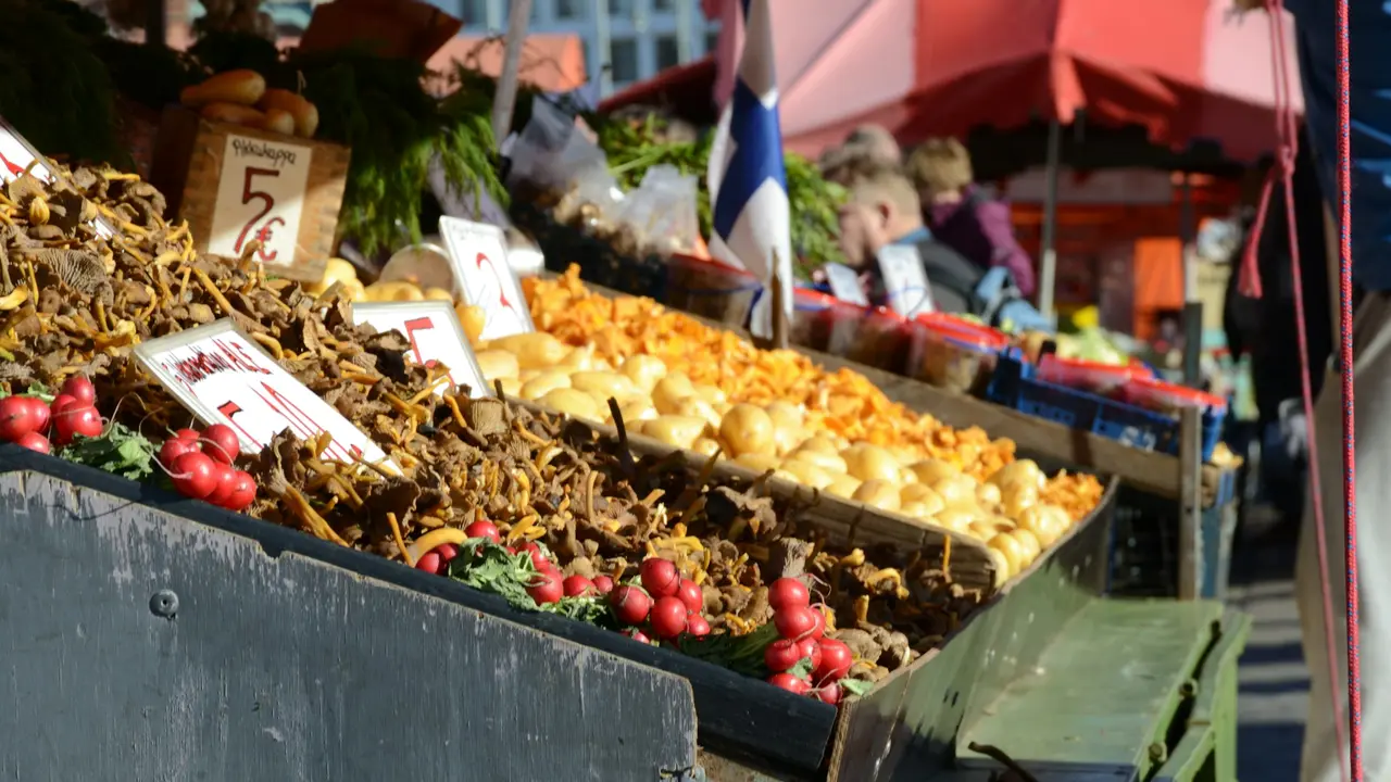Colorful produce and wild mushroom stalls at Kauppatori Market Square in Helsinki, Finland, featuring fresh chanterelles, potatoes, radishes, and local vendors on a sunny day.