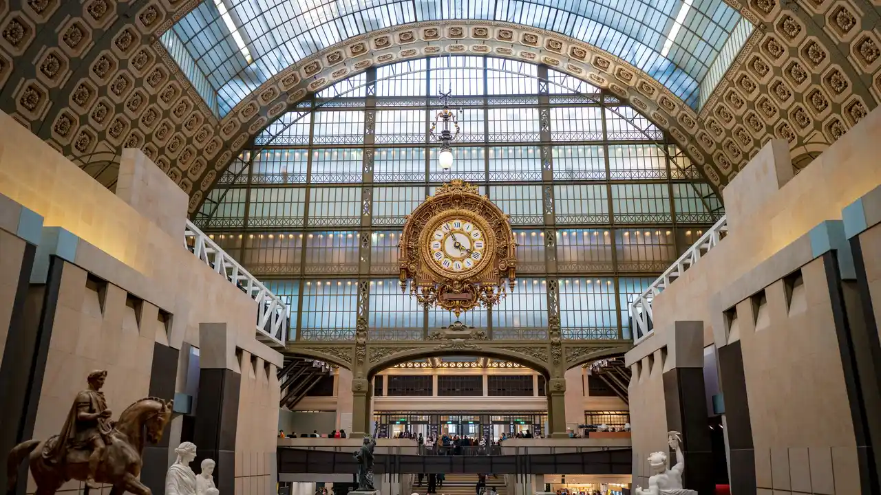 Interior of the Musée d’Orsay in Paris featuring the iconic large golden clock, grand glass-and-iron arched ceiling, and spacious museum hall with sculptures and visitors below.