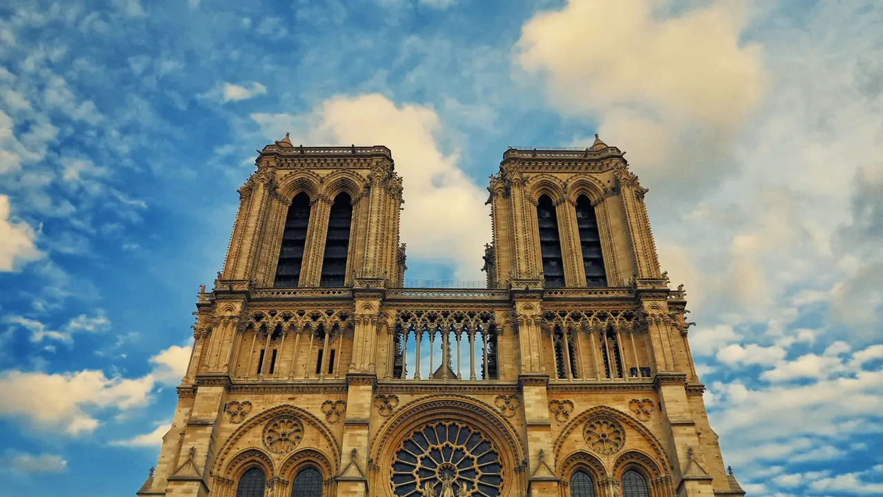 Notre-Dame Cathedral in Paris showcasing its iconic twin bell towers, Gothic architecture, and detailed stone façade against a blue sky with clouds.