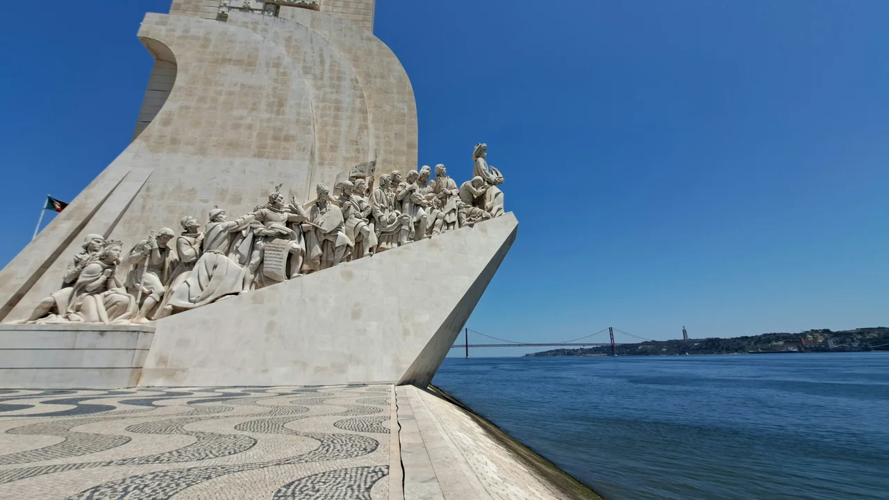 Monument to the Discoveries in Belém, Lisbon, featuring detailed stone sculptures of Portuguese explorers overlooking the Tagus River, with the 25 de Abril Bridge in the background on a clear sunny day.