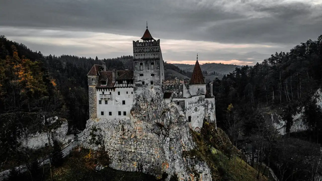 Bran Castle in Transylvania Romania perched on a rocky cliff at sunset surrounded by autumn forest under dramatic cloudy sky