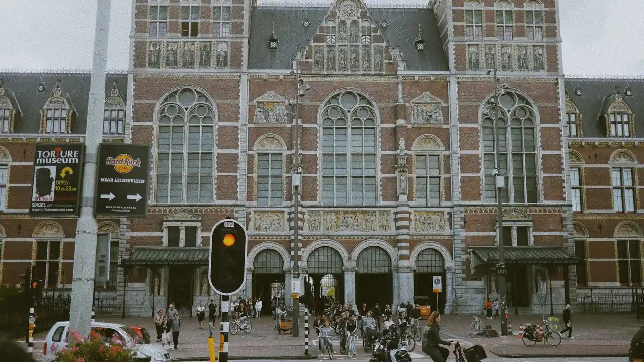 Exterior of the Rijksmuseum in Amsterdam with its historic Gothic Revival facade, arched entrances, and cyclists and pedestrians passing by on a busy city street.
