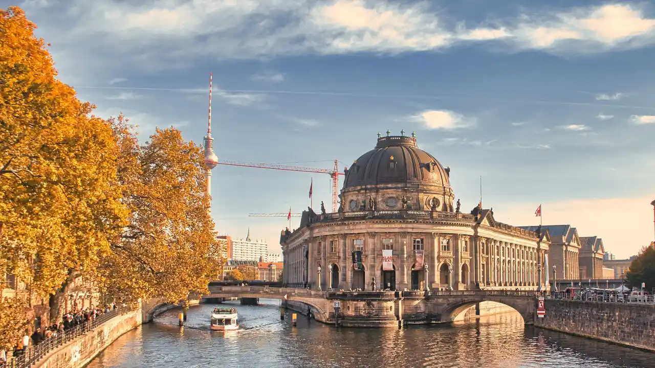 Scenic view of Museum Island in Berlin featuring the Bode Museum along the Spree River, autumn trees, boat traffic, and the Berlin TV Tower in the background under a clear sky.