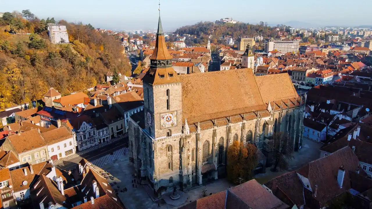 Aerial view of the Black Church (Biserica Neagră) in Brașov, Romania, surrounded by red-tiled rooftops and autumn hills in the historic Transylvania city center.