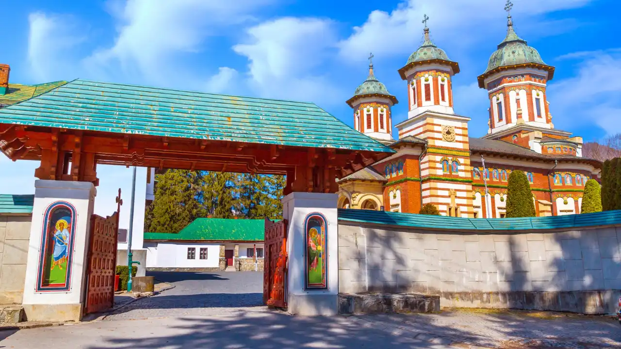 Colorful Romanian Orthodox monastery with three domed towers and painted entrance gate, featuring striped brick facade and religious icon murals under a bright blue sky.