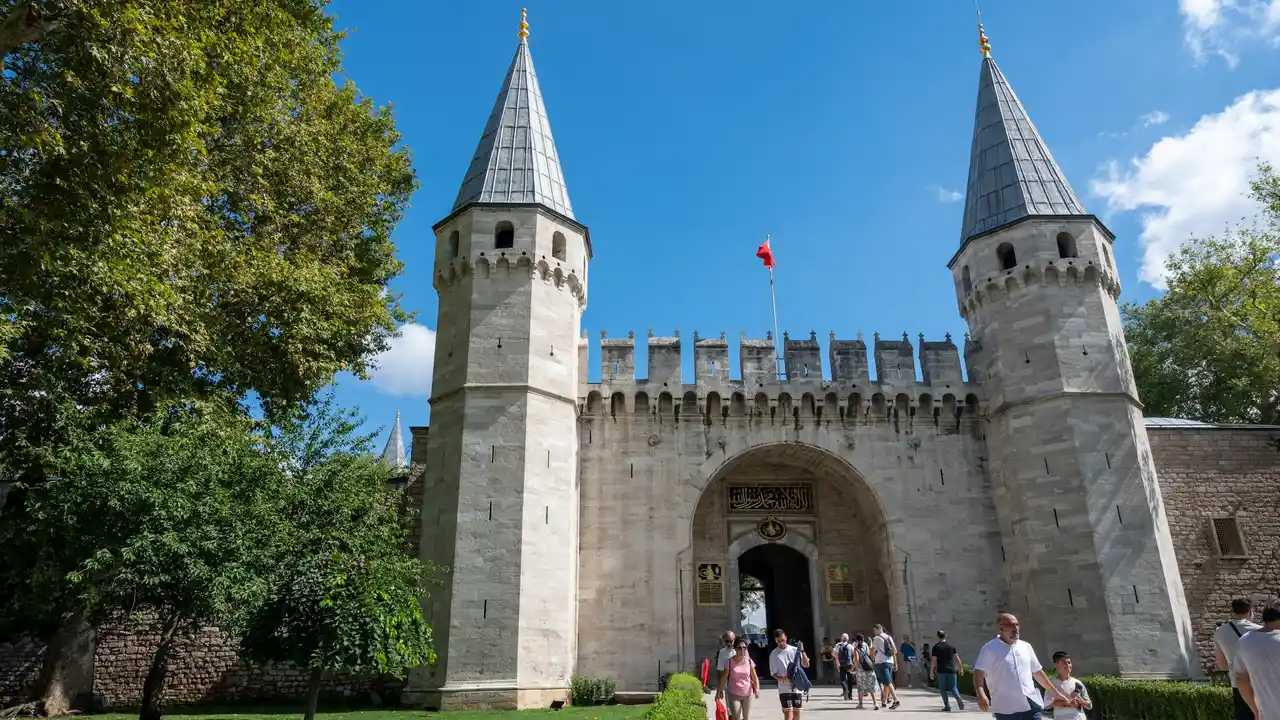 Visitors entering the Imperial Gate of Topkapi Palace in Istanbul, Turkey, featuring historic Ottoman architecture with twin stone towers, arched entrance, and Turkish flag under a bright blue sky