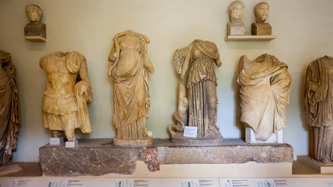 Ancient Greek and Roman marble statues displayed in a museum gallery, featuring headless classical sculptures with detailed drapery and busts mounted on a wall shelf.