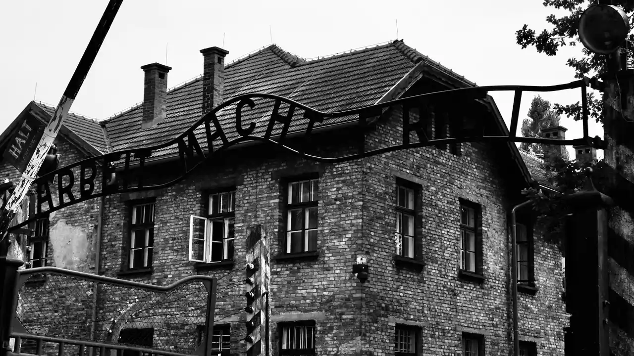 Entrance gate of the Auschwitz-Birkenau Memorial and Museum in Oświęcim, Poland, featuring the historic brick barracks and the “Arbeit Macht Frei” sign in a black-and-white photograph.
