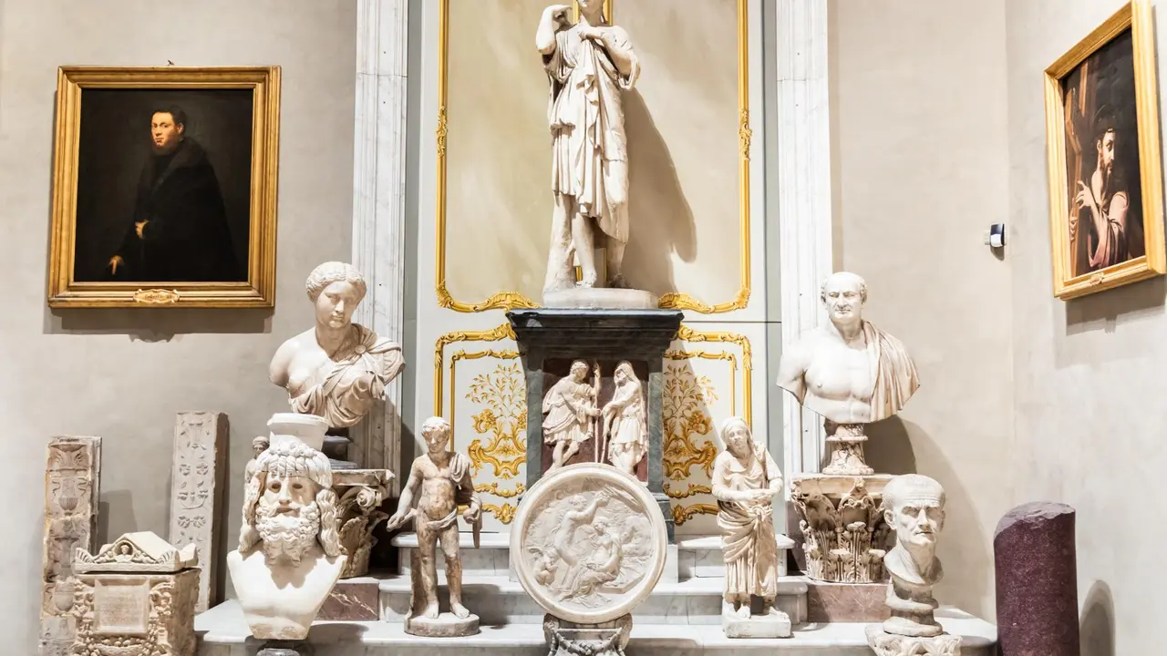 Ornate museum gallery interior featuring classical marble statues and busts displayed on a decorative altar, surrounded by framed Renaissance-style portraits on elegant cream and gold walls.