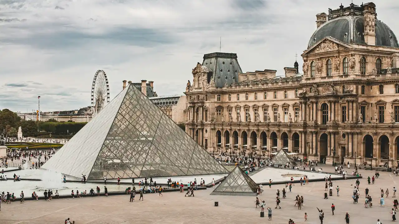 The Louvre Museum in Paris featuring the iconic glass pyramid in the main courtyard, with historic palace architecture and visitors walking around on a cloudy day.