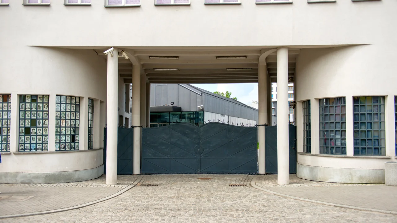 Entrance gate of a historic prison memorial site with guarded metal doors, memorial photo displays in windows, and institutional building architecture in an urban setting.