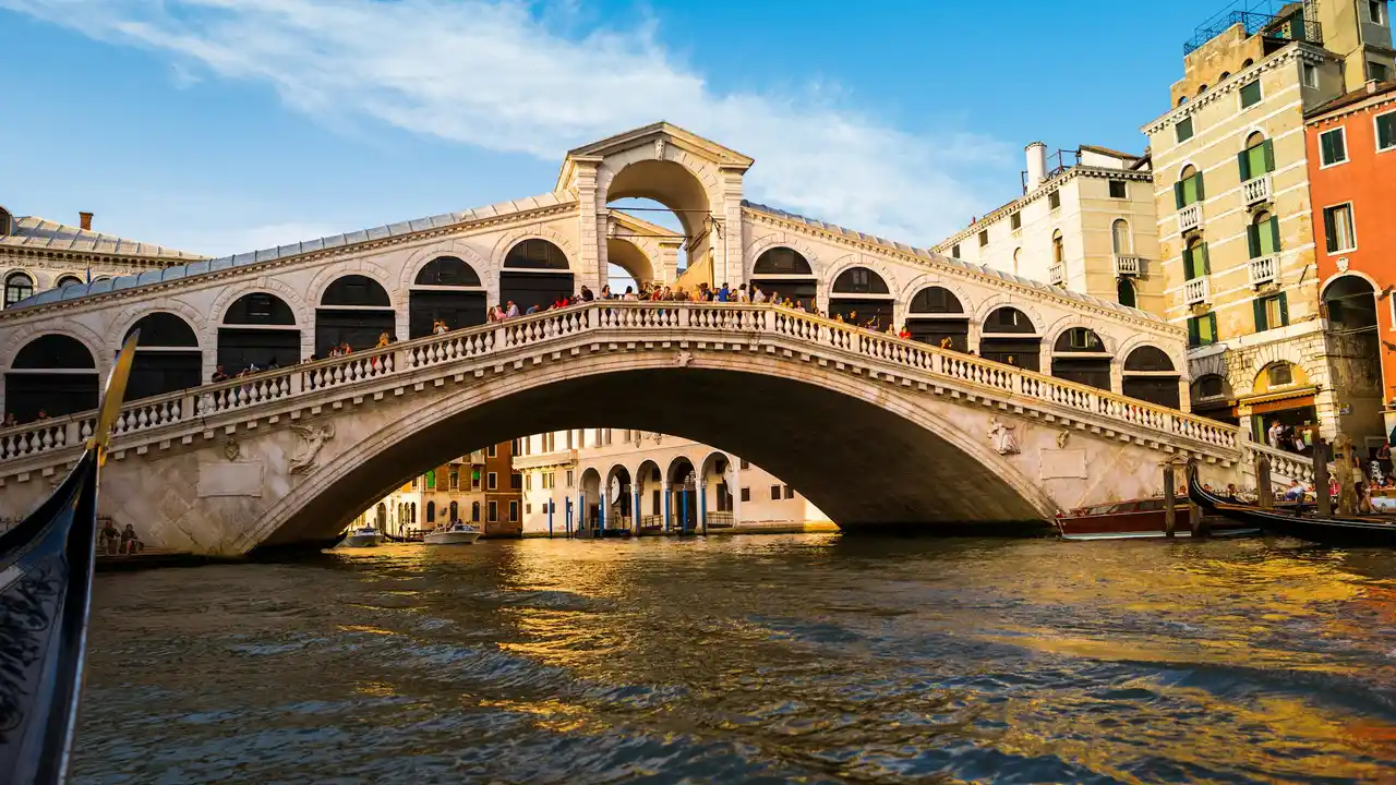 Rialto Bridge (Ponte di Rialto) spanning the Grand Canal in Venice, Italy, with historic arches, bustling pedestrians, gondolas, and colorful buildings along the water.