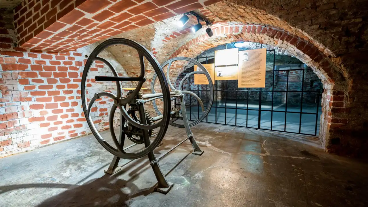 Historic industrial printing press machine displayed in a brick-arched museum basement, featuring vintage metal gears and wheels in a restored heritage exhibition space