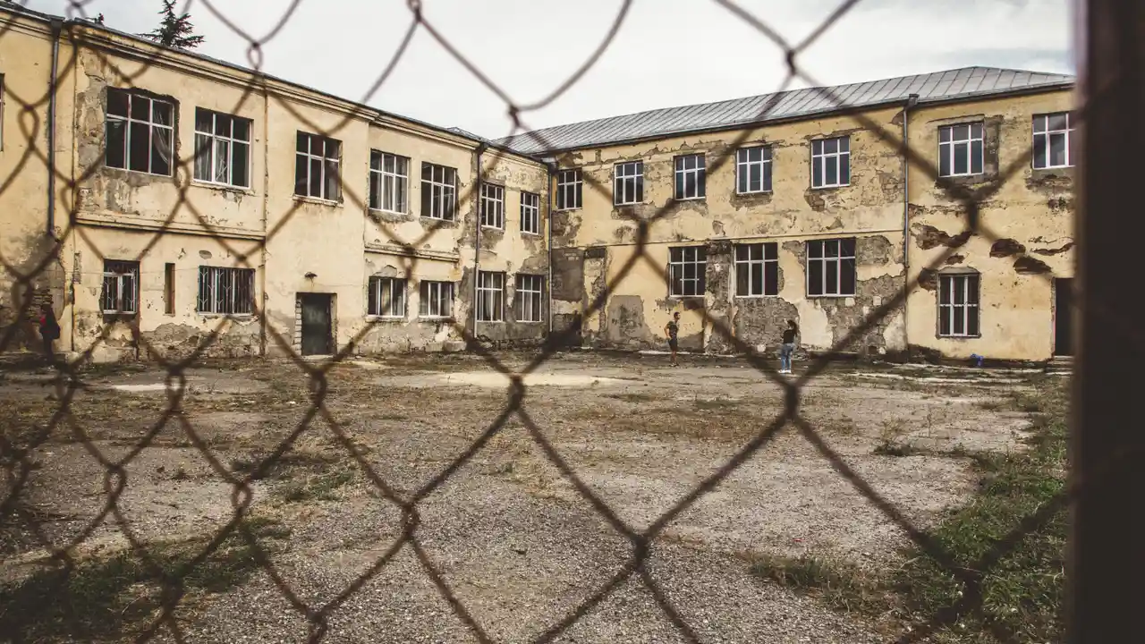 Abandoned, dilapidated building viewed through chain-link fence, featuring broken windows, peeling paint, and cracked walls in an empty courtyard, symbolizing urban decay and neglect