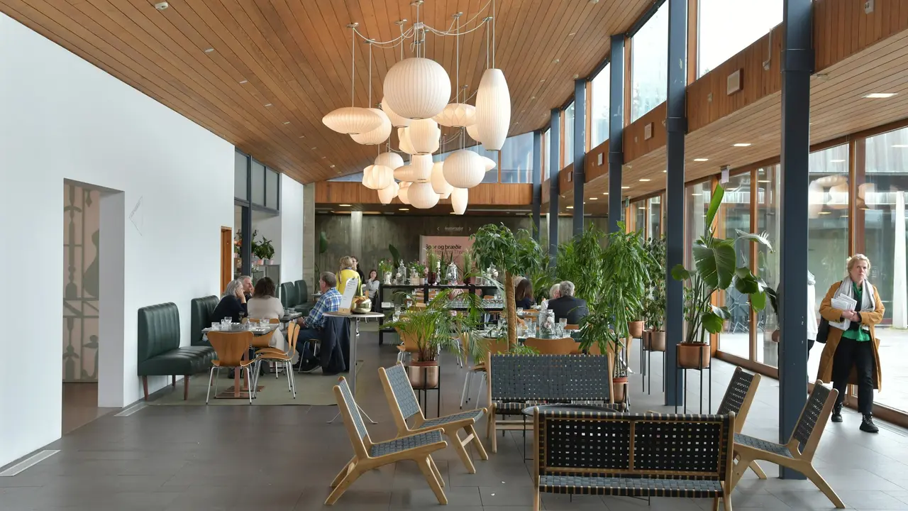 A brightly lit, modern museum café with floor-to-ceiling glass windows, wood-paneled ceilings, and large white spherical pendant lights hanging over patrons seated at various tables and lounge chairs.