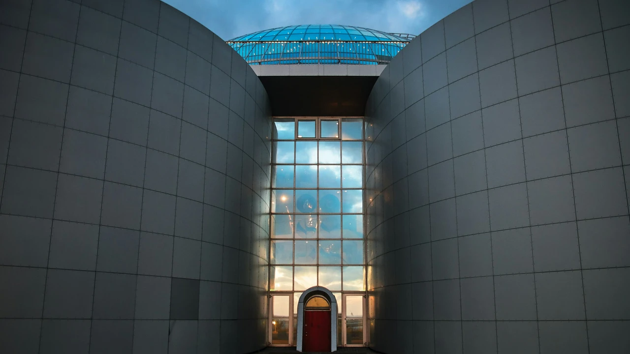 A symmetrical, low-angle shot of the modern Perlan Museum in Reykjavík, featuring two large, curved grey concrete walls flanking a tall glass-windowed entryway that reflects the blue sky, topped with a distinctive blue glass dome.