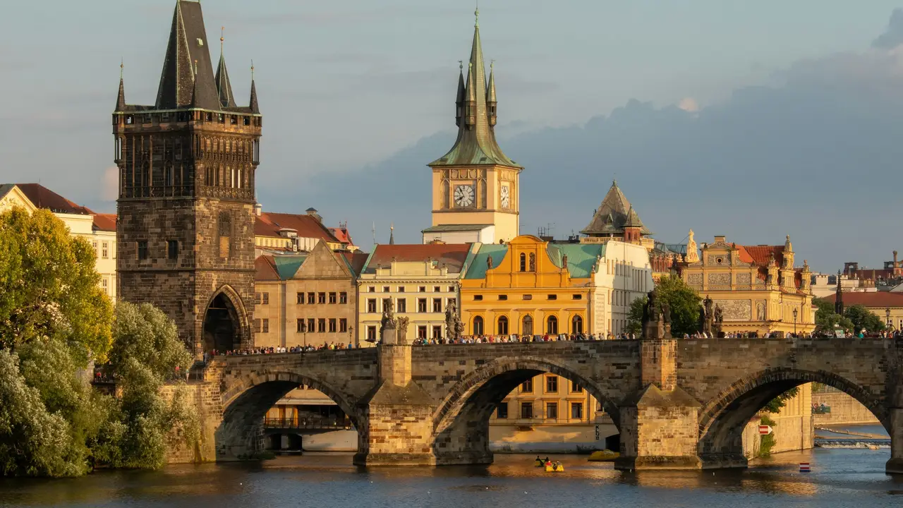A close-up view of the historic Charles Bridge in Prague during the golden hour. The image features the ornate, dark stone Old Town Bridge Tower at the bridge's entrance, with the stone arches of the bridge extending across the Vltava River.