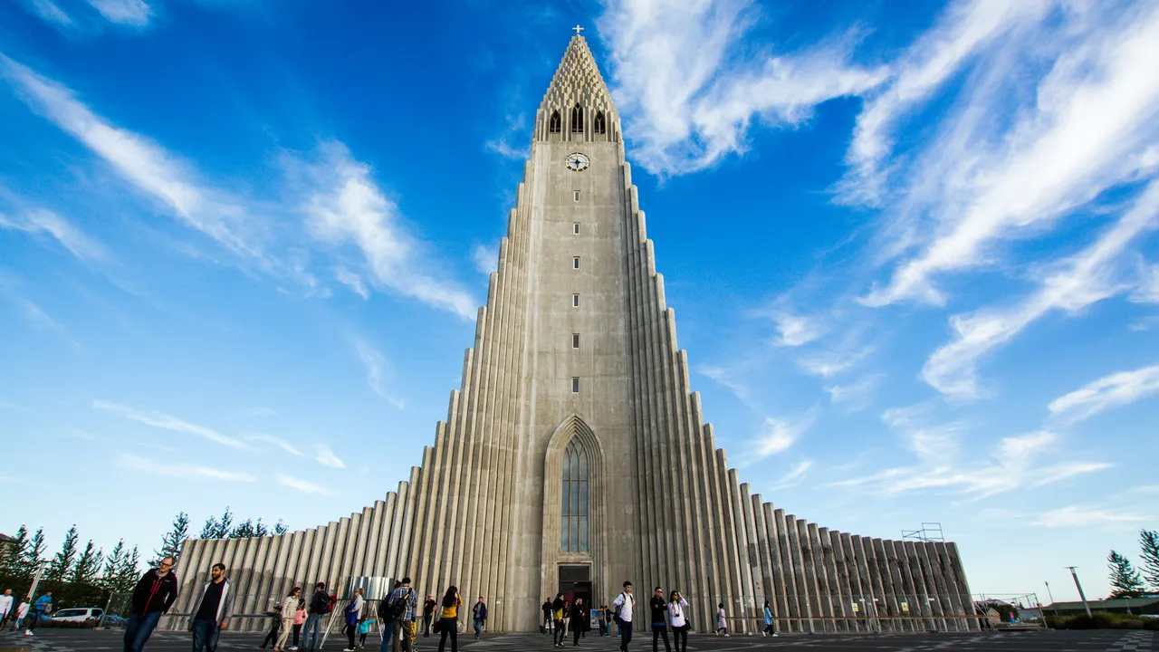 A wide-angle, low-elevation shot of Hallgrímskirkja church in Reykjavík, Iceland, featuring its unique stepped concrete facade and central bell tower rising into a bright blue sky with wispy clouds, while people walk across the large plaza in front.