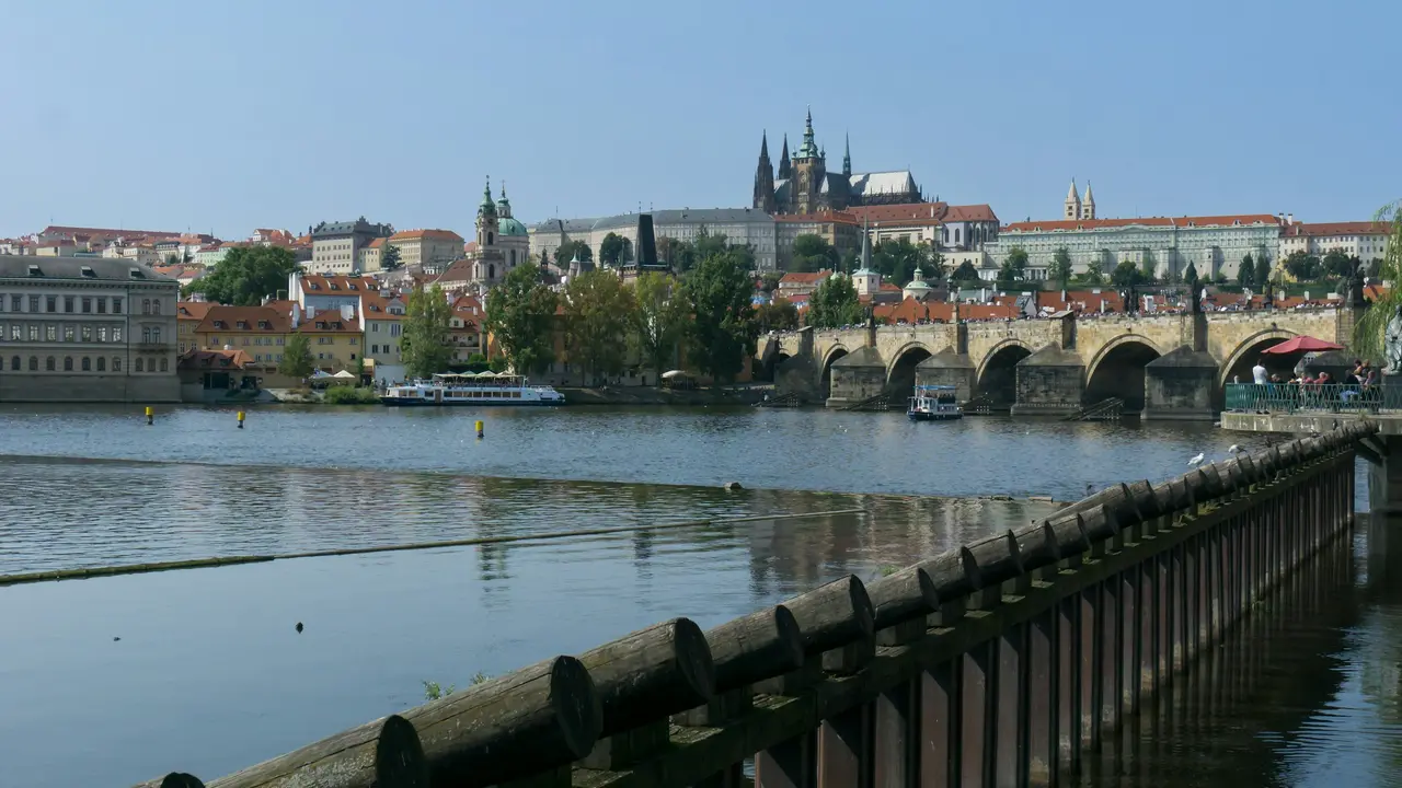 A wide-angle, scenic view of the Prague skyline under a bright blue sky. The image features the historic Charles Bridge with its stone arches spanning the Vltava River, where small boats are visible. In the background, Prague Castle and the spires of St. Vitus Cathedral dominate the hilltop, surrounded by the city's signature red-tiled roofs and greenery.