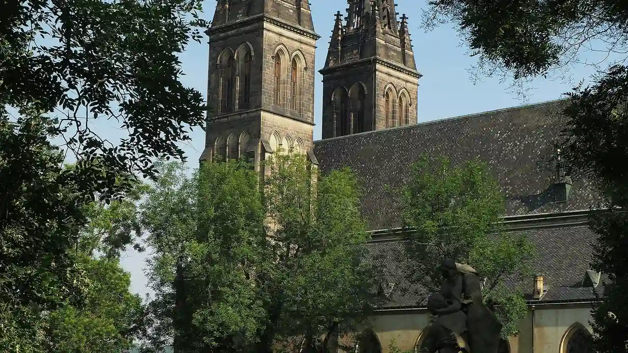 A view of the Basilica of St. Peter and St. Paul at Vyšehrad in Prague, framed by lush green trees. The image captures the two prominent neo-Gothic stone towers and the dark tiled roof of the church against a clear, light blue sky. In the lower foreground, a dark stone statue is partially visible among the foliage.