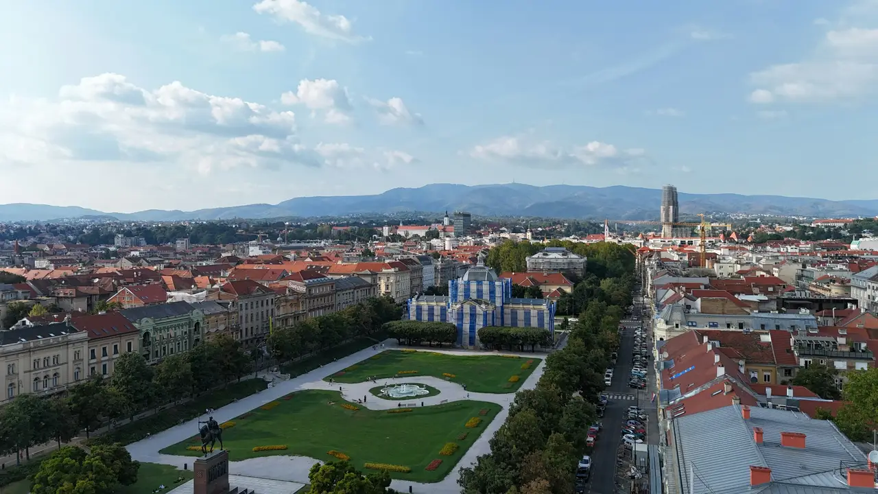 An aerial, high-angle view of King Tomislav Square in Zagreb, Croatia, showcasing a large, green rectangular park with manicured lawns and a central fountain. In the foreground, a tall bronze equestrian statue stands on a stone pedestal, while the blue-scaffolded Art Pavilion sits at the far end of the square.