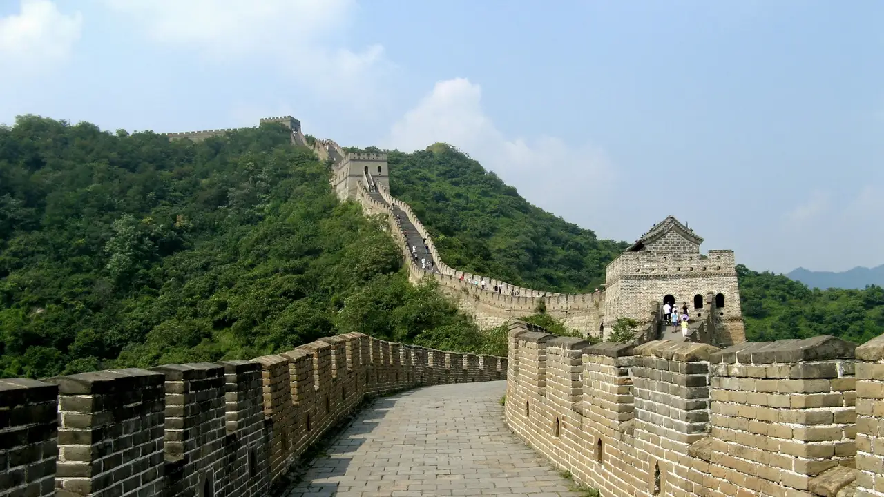 A wide-angle, eye-level shot of the Great Wall of China snaking up a steep, lush green mountain under a clear blue sky. In the foreground, a stone-paved walkway leads toward a sturdy watchtower with arched windows, while further up the ridge, another watchtower sits at a higher elevation. Small figures of tourists can be seen walking along the undulating path of the wall as it disappears into the distant hills.