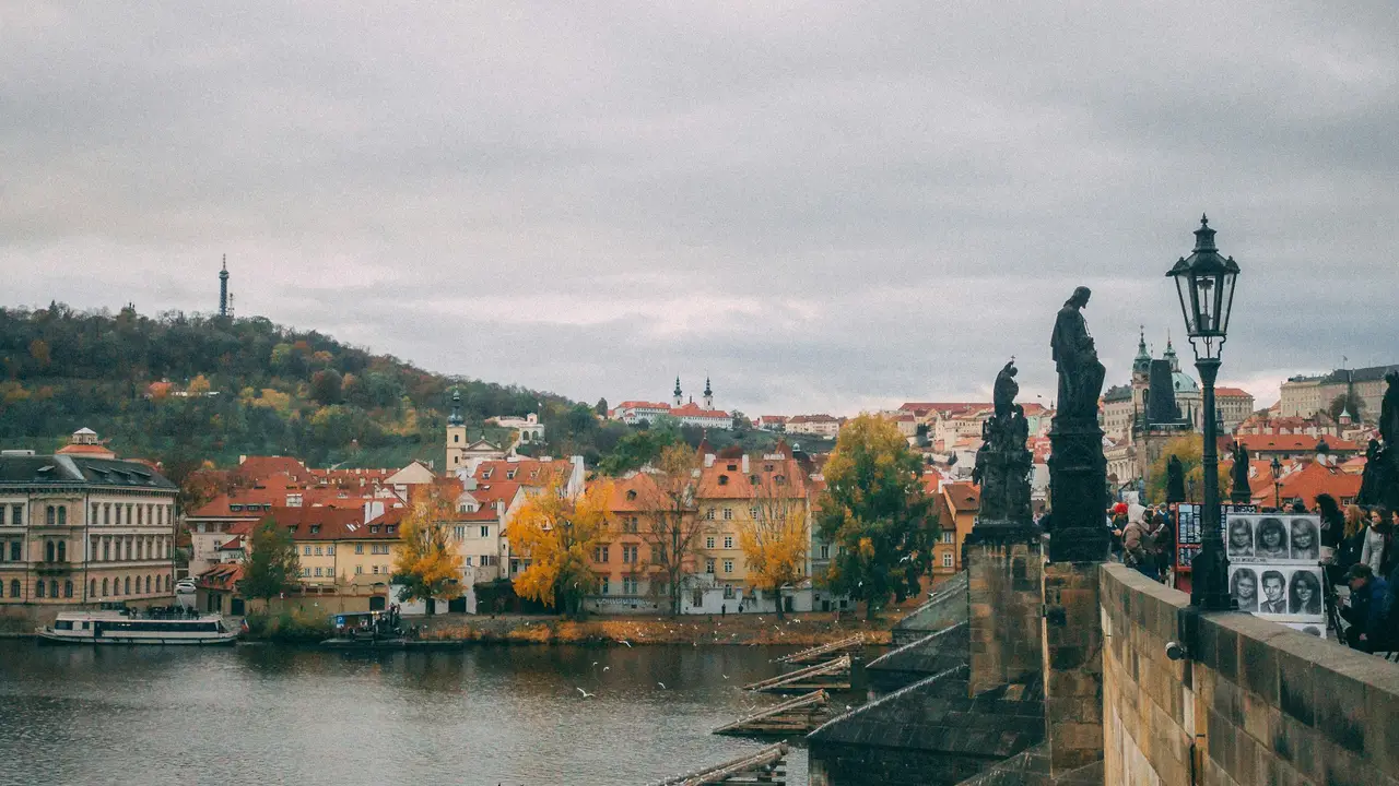 view from the stone parapet of the Charles Bridge in Prague, looking across the Vltava River toward the Lesser Town (Malá Strana). The scene is captured on an overcast day, highlighting the red-tiled roofs of historic buildings and trees with yellow autumn foliage along the riverbank. In the distance, the Petřín Lookout Tower stands atop a lush green hill.