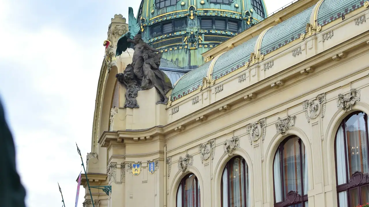 A close-up, low-angle view of the Municipal House in Prague, focusing on its ornate Art Nouveau architecture. The image highlights a large, green-patina dome with gold accents and a detailed stone sculpture of a winged figure perched on the building's facade. The cream-colored exterior is decorated with intricate carvings, shields, and large arched windows under a pale, overcast sky.