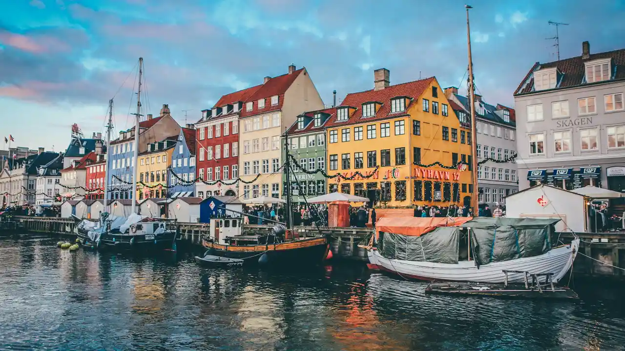 A wide-angle, eye-level shot of the Nyhavn canal in Copenhagen at twilight, featuring traditional wooden boats docked along the water. The canal is lined with iconic, brightly colored historic townhouses in shades of yellow, red, and blue, some adorned with festive string lights. The sky above is a mix of soft blue and pink clouds, which are reflected in the rippling surface of the water.