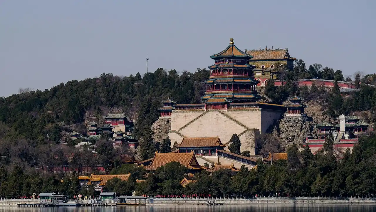 A wide-angle, low-angle shot of the Tower of Buddhist Incense at the Summer Palace in Beijing, perched majestically on Longevity Hill. The multi-tiered, octagonal pagoda features traditional blue-tiled roofs and red columns, rising above a massive stone base and surrounded by dense green trees. In the foreground, the calm water of Kunming Lake reflects the clear sky, with a small dock visible along the shoreline.