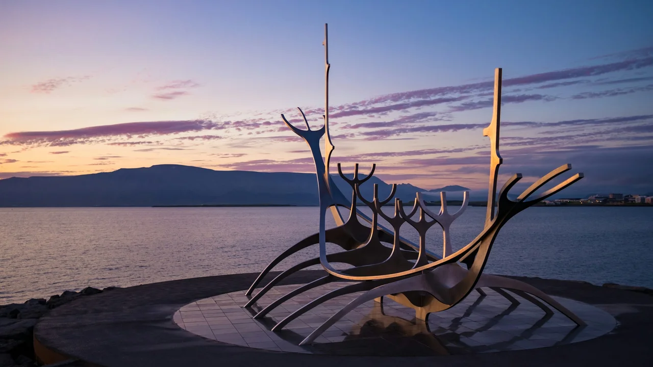 A sleek, stainless steel sculpture of a stylized Viking longboat, known as the Sun Voyager (Sólfar), sits on a circular granite platform at the water's edge in Reykjavík, Iceland, set against a calm bay and a purple-streaked sunset sky.