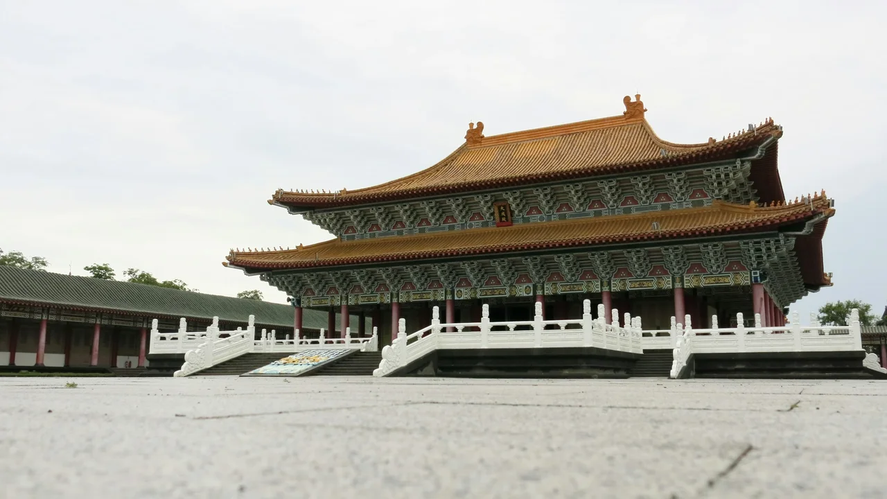 A wide-angle, low-angle shot of a grand, traditional Chinese hall at the Temple of Confucius. The building features a multi-tiered, golden-yellow tiled roof with ornate green and red eaves, supported by a row of thick red columns. A wide, grey stone courtyard leads to the structure, which sits on a raised platform enclosed by a decorative white marble balustrade and sets of stairs. The sky is a soft, overcast grey.