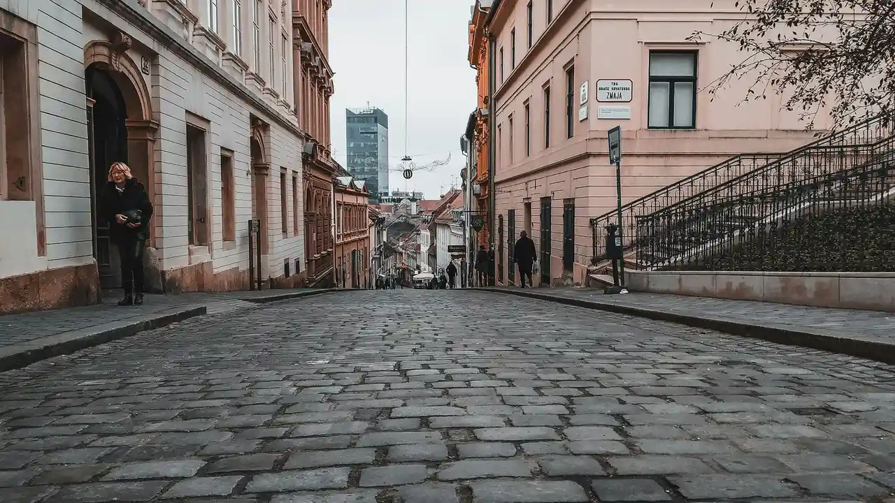 A wide-angle, low-angle shot of a cobblestone street in the Upper Town of Zagreb, Croatia, sloping steeply downward toward a misty city center. Historic multi-story buildings in shades of cream and pink line the street, including one with a stone archway where a person stands in the shadows. To the right, a set of stone stairs with a decorative wrought-iron railing leads upward, while in the far distance, a modern glass skyscraper rises above the traditional rooftops under an overcast sky.