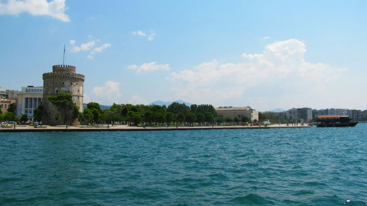 White Tower of Thessaloniki standing along the waterfront promenade, with blue sea in the foreground and city buildings under a clear sky.